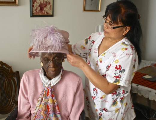 A caregiver in a patterned scrub top helps an elderly woman put on a pink hat adorned with feathers. The elderly woman is seated, wearing glasses, a pink sweater, and a colorful scarf. The setting appears to be a home with framed pictures on the wall and a table in the background.