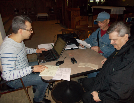 Three men sitting around a table. One man is working on a laptop while the other two are sitting next to him, looking at some documents. One man is wearing a blue cap and a jacket, while the other man is wearing a black jacket. The background features a church-like interior with wooden furniture. The scene appears to be a meeting or discussion involving paperwork.