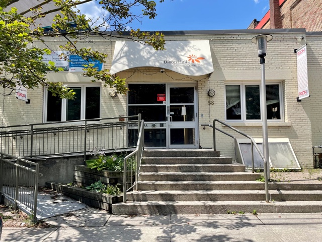 The exterior of a building with the sign "Dixon Hall" displayed above the entrance. The building has a ramp for accessibility and a set of stairs leading up to the front door. There are a few small plants and greenery around the entrance, and a couple of signs are visible next to the building. The sky is clear, and the scene is brightly lit by sunlight.