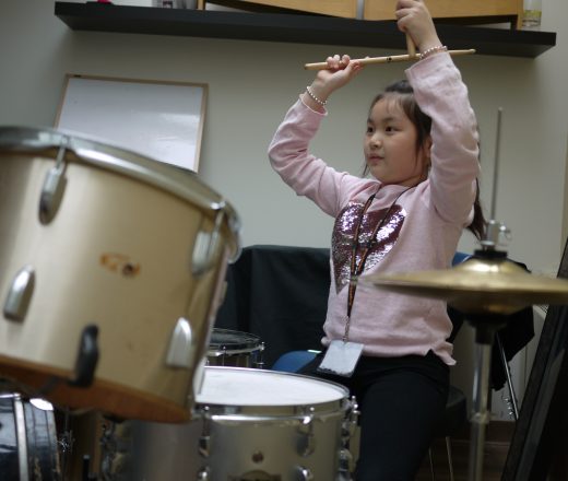 A young girl wearing a pink sweater with a sequined heart design is playing a drum set. She is holding drumsticks high above her head, preparing to strike the drums. The setting appears to be a music room or practice space, with a whiteboard and shelves in the background.