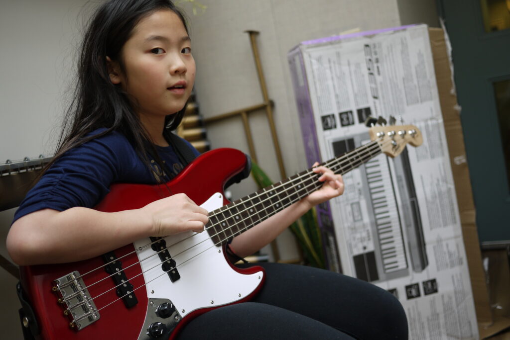 A young girl with long dark hair is playing a red and white bass guitar. She is seated and looking towards the camera with a focused expression. The background includes a large cardboard box with a keyboard image on it and some musical equipment.