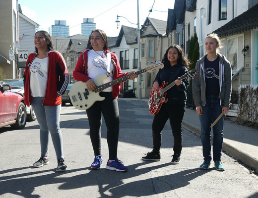 Four young musicians stand together on a residential street in daylight. Two of them are holding guitars—one with a white bass guitar and another with a red electric guitar—while another holds drumsticks. They are dressed casually, with matching shirts featuring a circular logo, and appear to be part of a band. The background features houses, parked cars, and city buildings in the distance.