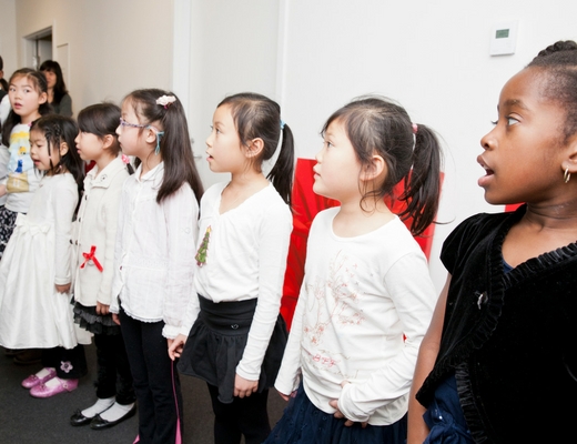 A group of young children dressed in formal attire stands in a line, singing or performing. They are looking in the same direction, appearing focused. The background shows a bright indoor setting with a white wall and a hallway.