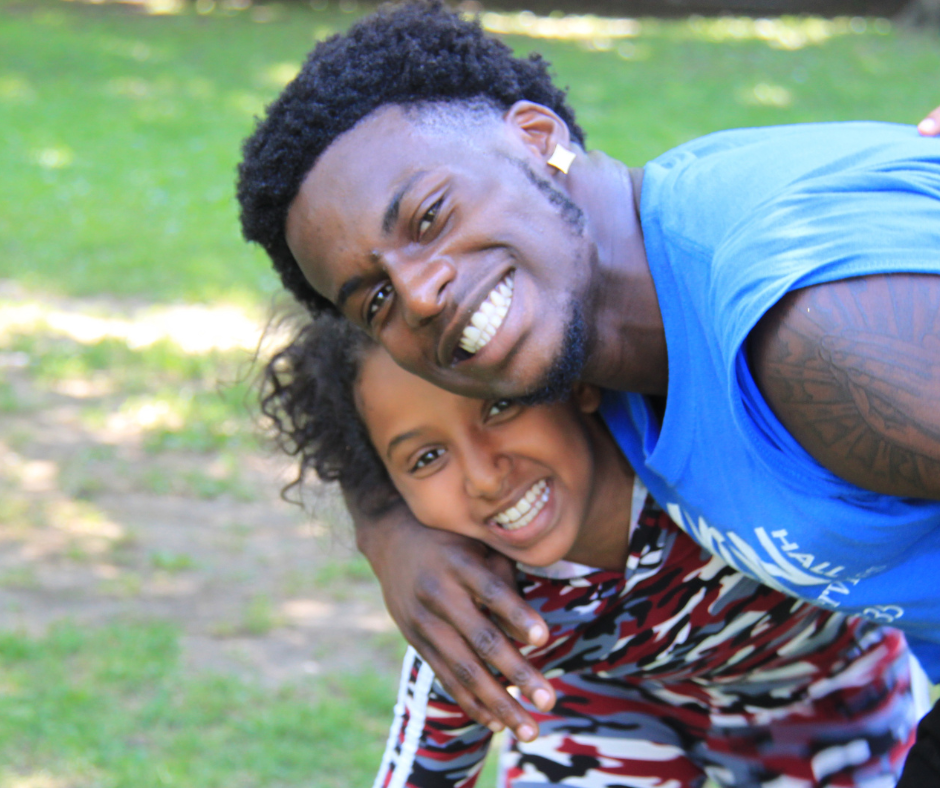 Untitled design (24) Two people smiling outdoors, with an adult in a blue shirt hugging a child in a patterned outfit. They are both looking at the camera. Grass is visible in the background.