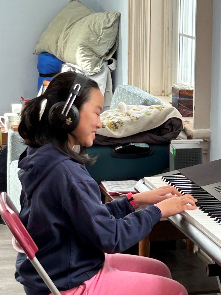 A young girl wearing a navy hoodie and pink pants is playing a digital keyboard while wearing large headphones. She is seated on a red folding chair, focused and smiling as she plays. The background includes a window with natural light and a stack of pillows and blankets.