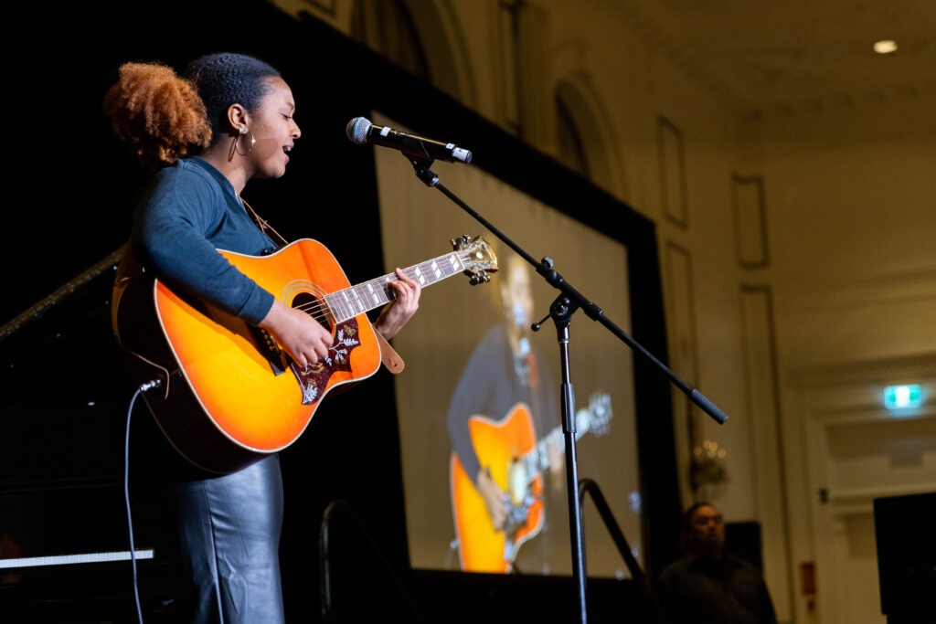 A singer-songwriter performs on stage with an acoustic guitar, singing into a microphone while a large screen behind her displays a live projection of her performance.