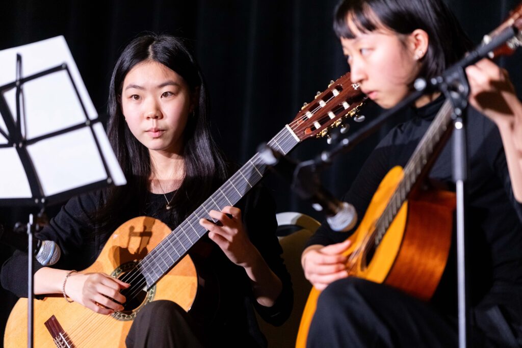 Two young women in black perform on classical guitars, reading from sheet music on a stand, with microphones capturing their music.