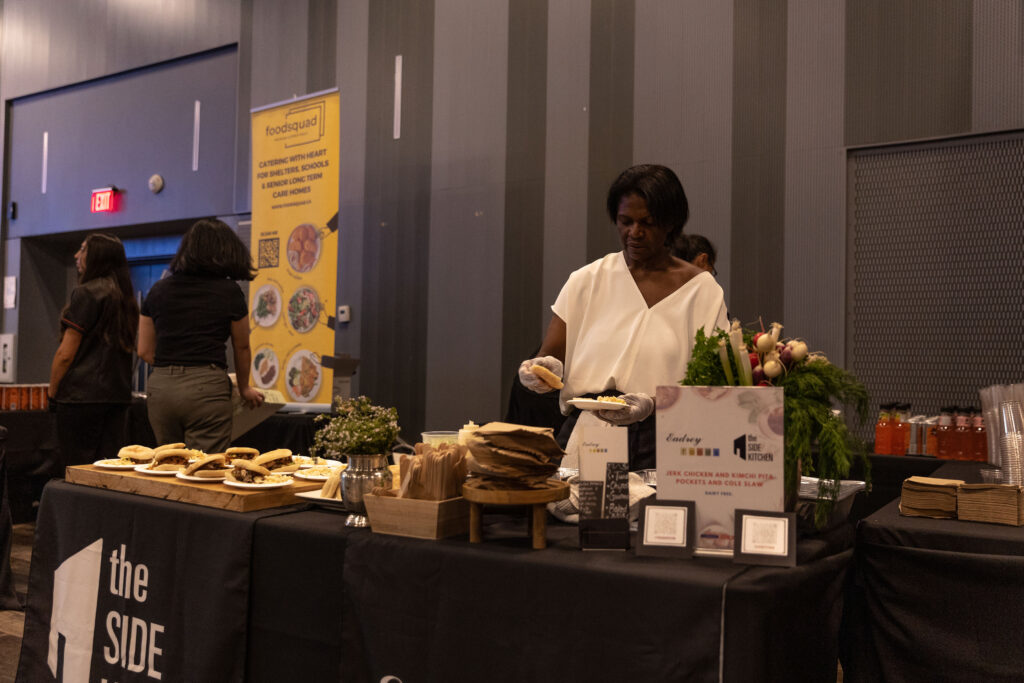 A woman in a white blouse serves food at a catering table filled with sandwiches, fresh ingredients, and promotional signage. A yellow "FoodSquad" banner is visible in the background.