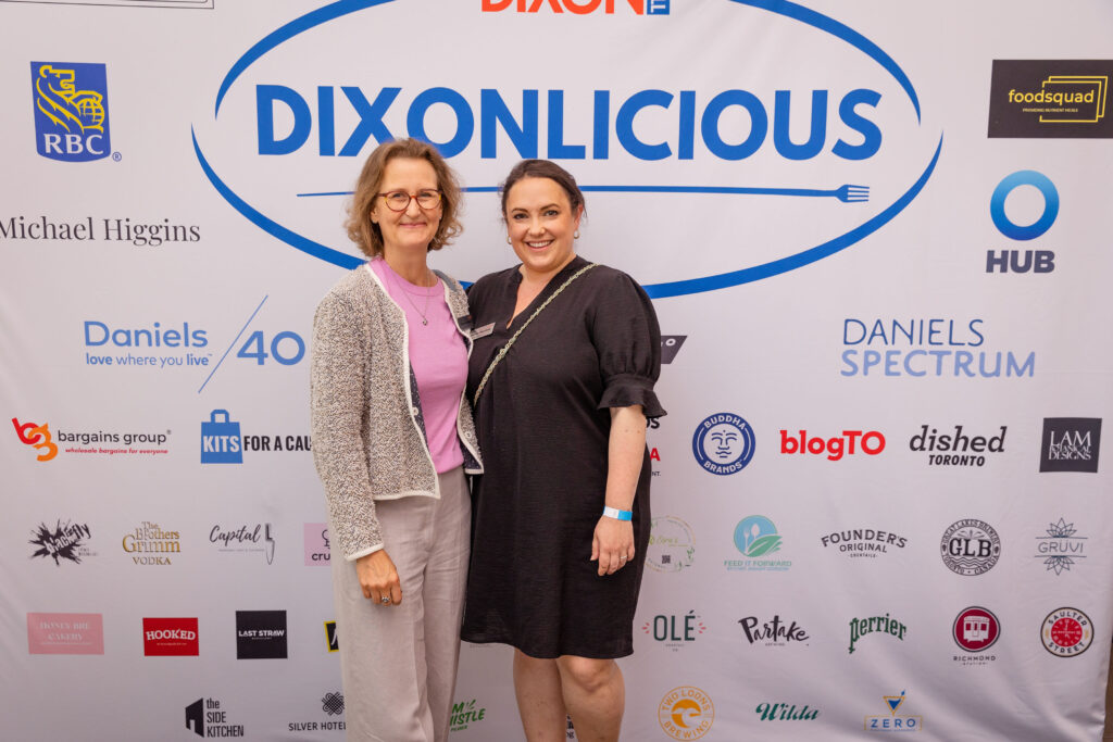 Two women smile and pose in front of a large "DIXONLICIOUS" event backdrop featuring sponsor logos.
