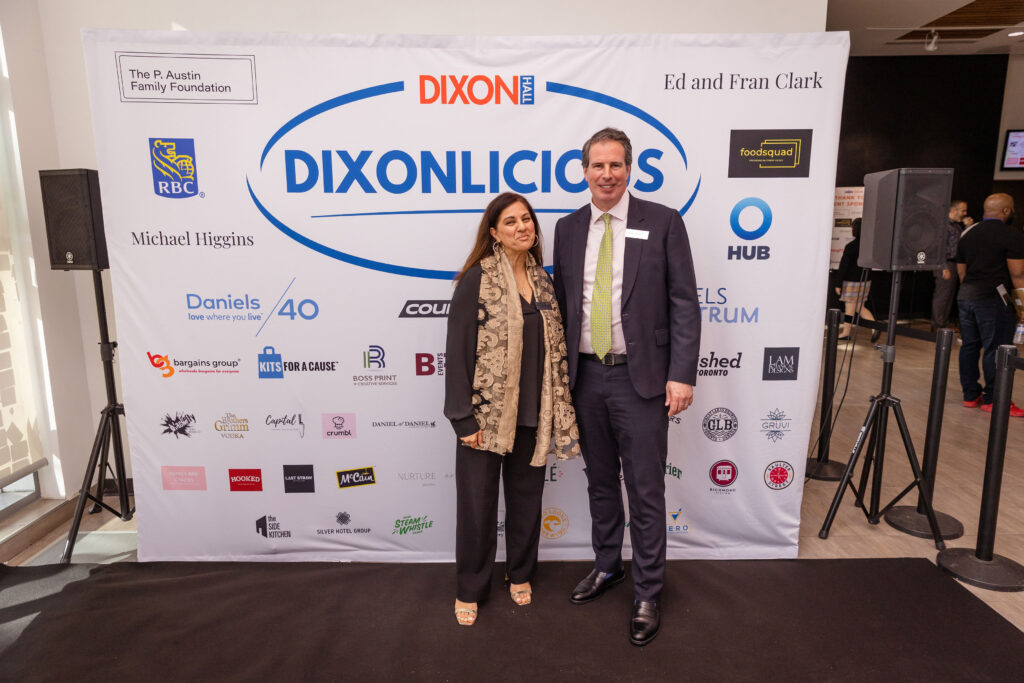 A man in a suit and a woman in a black outfit with a patterned scarf pose in front of a "DIXONLICIOUS" event backdrop featuring various sponsor logos.