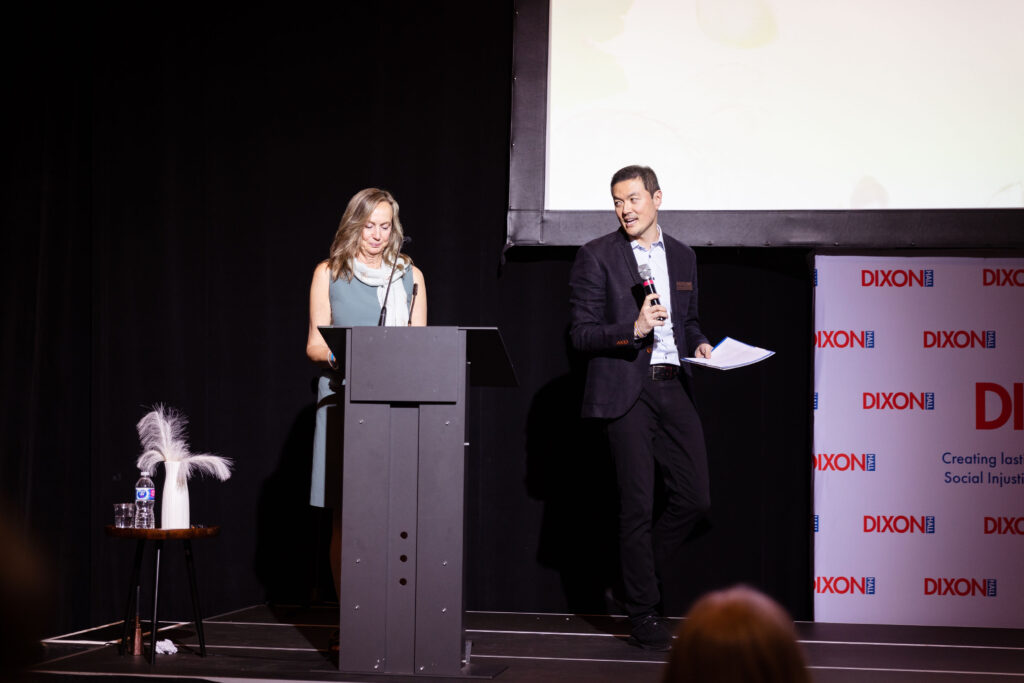 A woman stands at a podium speaking while a man beside her holds a microphone and papers, addressing an audience at an event.
