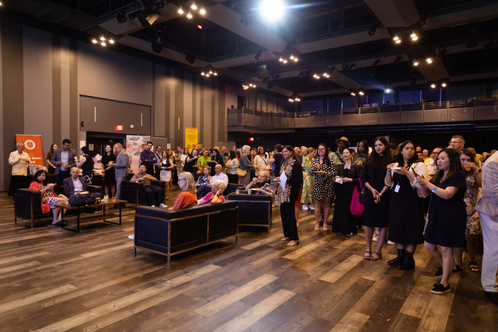 A large crowd of people gathered in an event space, some standing and some seated on couches, attentively watching a presentation.