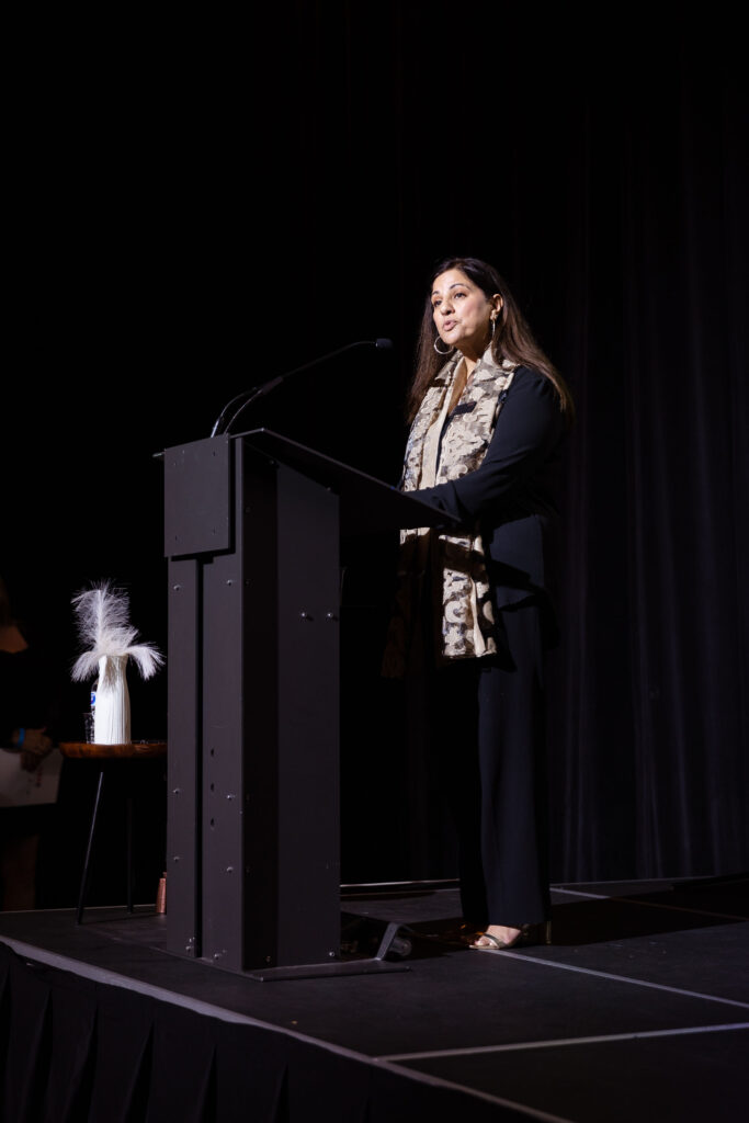 A woman in a black outfit with a patterned scarf speaks at a podium on stage.