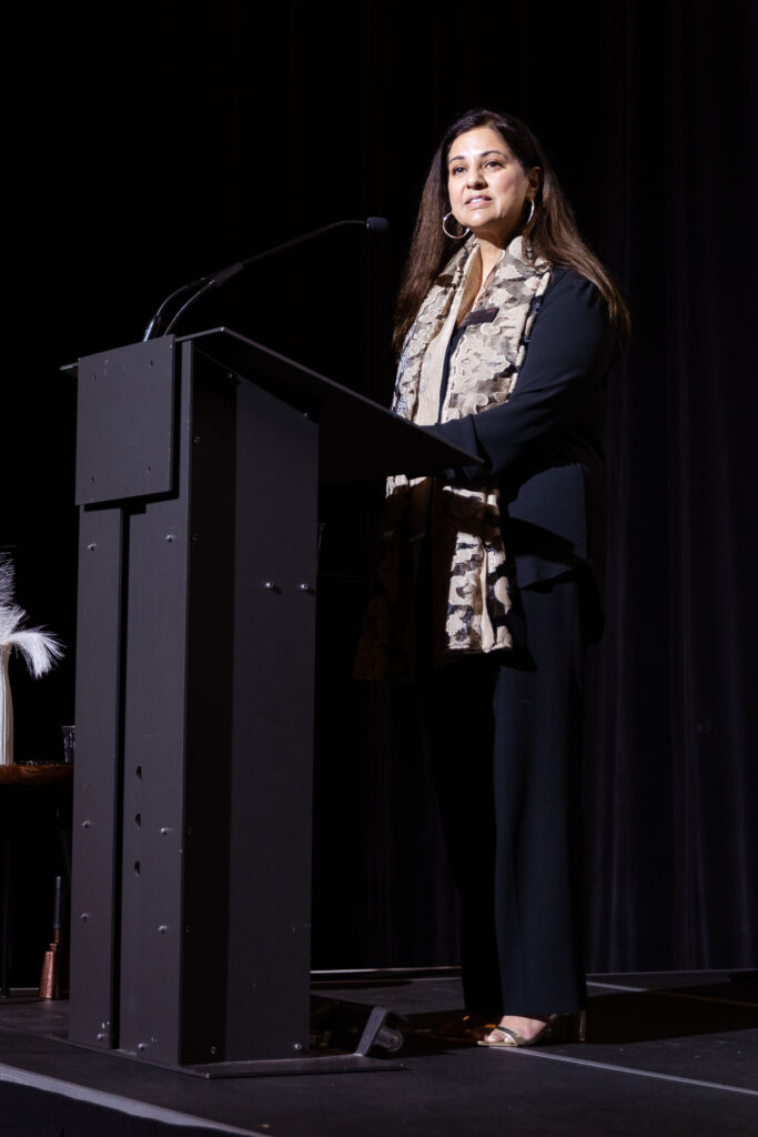 A woman in a black outfit with a patterned scarf speaks at a podium on stage.