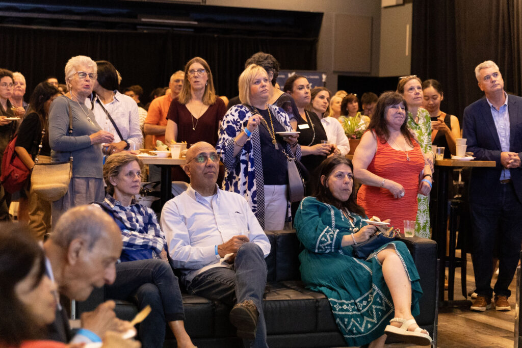 A group of people, some seated and some standing, attentively watching an event while enjoying food and drinks.