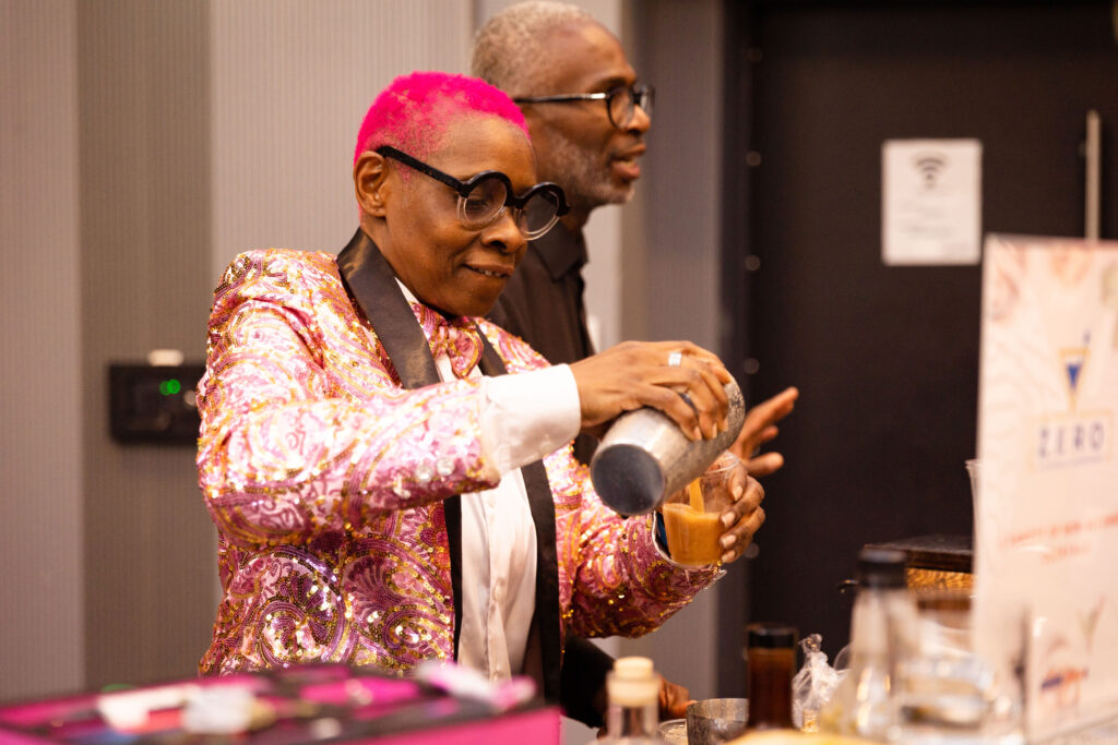 A bartender with bright pink hair and a shiny patterned jacket pours a drink from a shaker, with another person standing behind them.