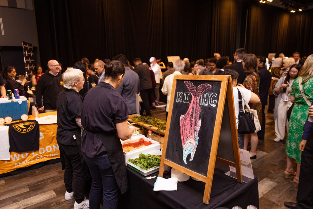 A food station with chefs preparing dishes, featuring a chalkboard sign with a drawing of a fish labeled "KING," and a crowd of attendees in the background.