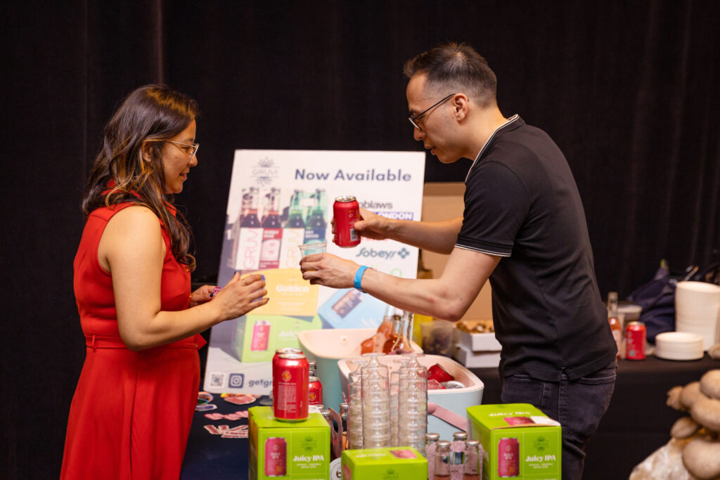 A vendor in a black shirt handing a canned beverage to a woman in a red dress at a tasting station, with promotional signage and product displays in the background.