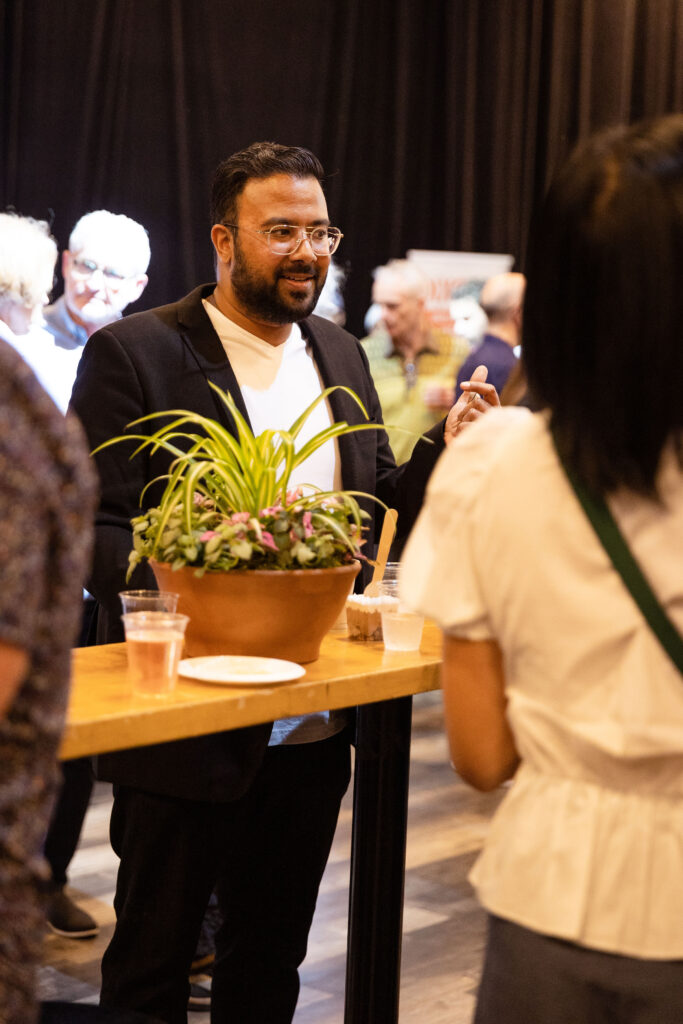 A man in a black blazer and white shirt is engaged in conversation with a woman at a high-top table with drinks and a potted plant, surrounded by other event attendees.