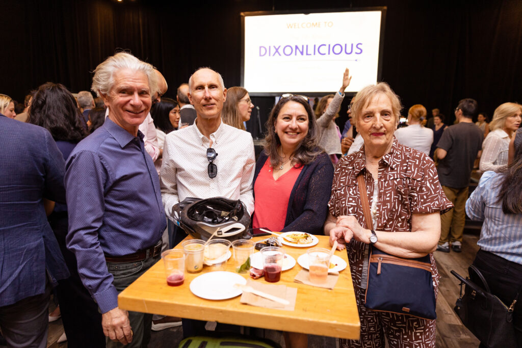 Four people pose together at an event, standing around a table with food and drinks. A large screen in the background displays "DIXONLICIOUS."