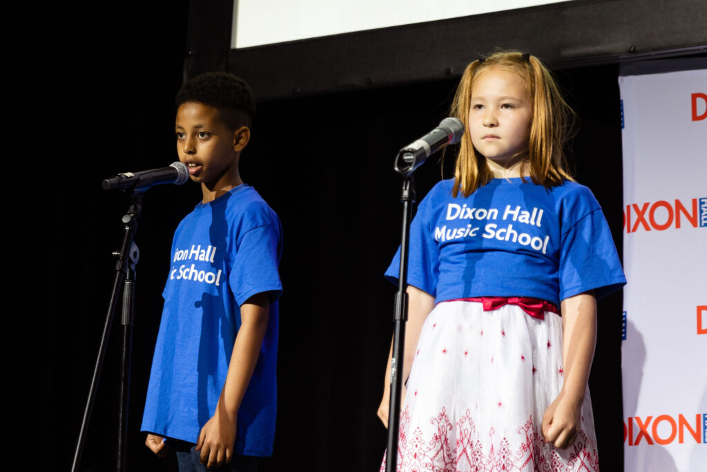 Two young children in blue "Dixon Hall Music School" shirts stand on stage, each in front of a microphone, performing.