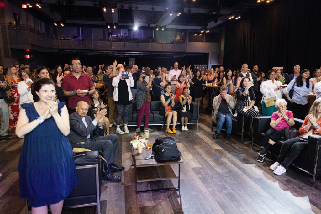 A large audience in an indoor venue claps and takes photos, showing enthusiasm and engagement.