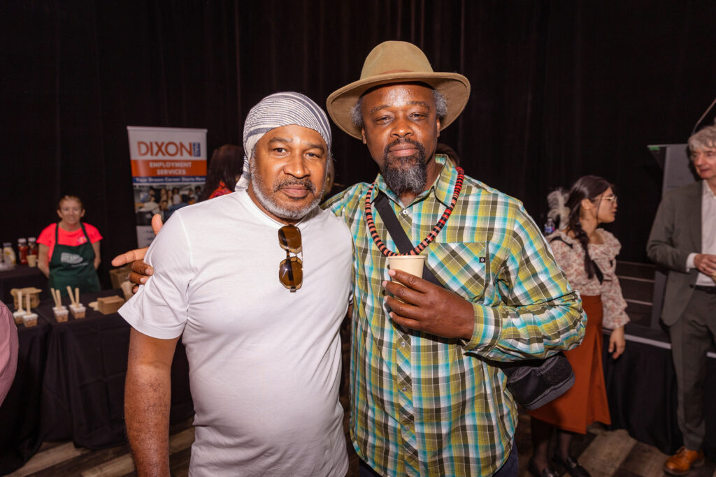 Two men pose together at an event, one wearing a white shirt and headscarf with sunglasses hanging from his shirt, and the other wearing a plaid shirt, beaded necklace, and a hat while holding a drink.
