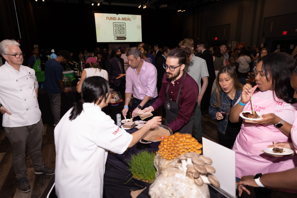 A chef in a white coat serves food to an attendee wearing an apron at a busy event. A table in the foreground displays fresh mushrooms and herbs. A crowd mingles in the background, with a "FUND-A-MEAL" sign projected on the screen.