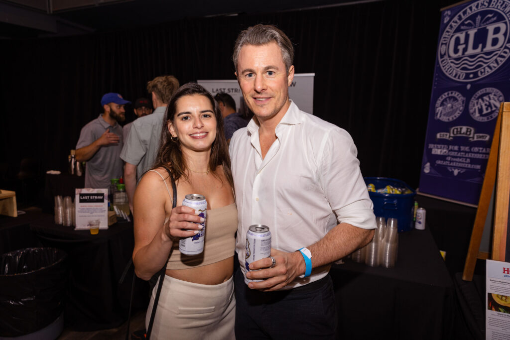 A smiling couple holding canned drinks at an event, surrounded by other attendees.
