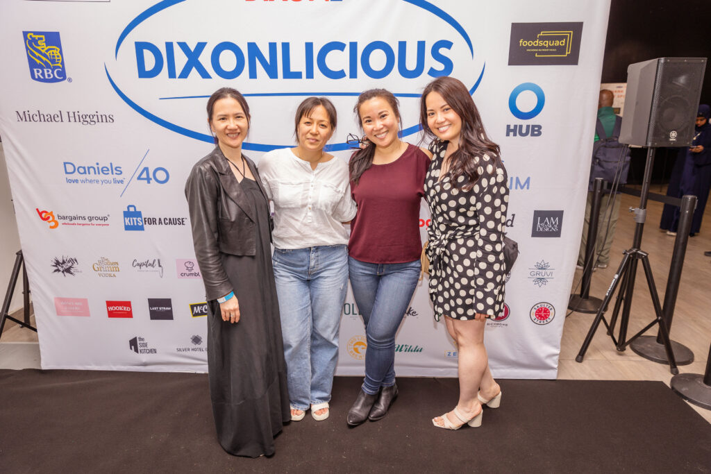 Four smiling women posing in front of a "DIXONLICIOUS" event backdrop featuring various sponsor logos.
