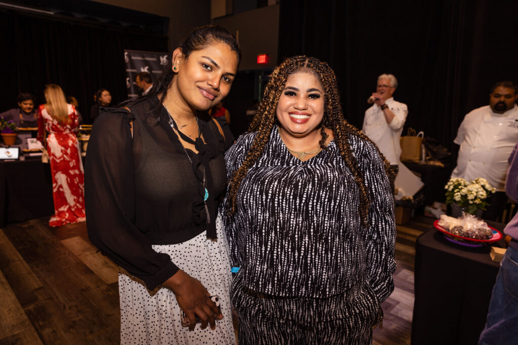 Two women smiling and posing together at an event, one wearing a black and white blouse with matching pants and the other in a black sheer top with a white skirt.
