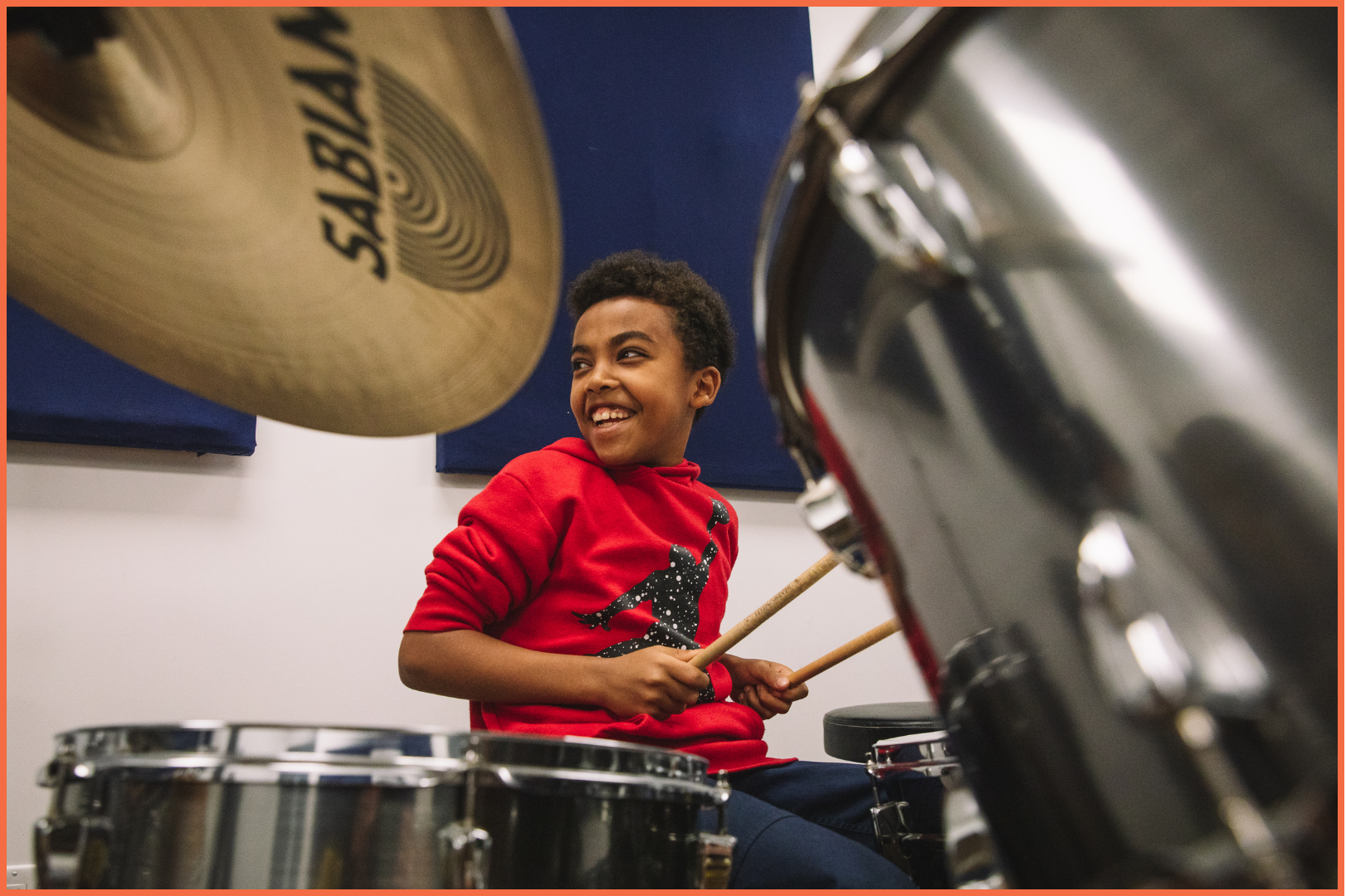A young boy in a red hoodie smiles while playing the drums in a music room, surrounded by cymbals and drum equipment.