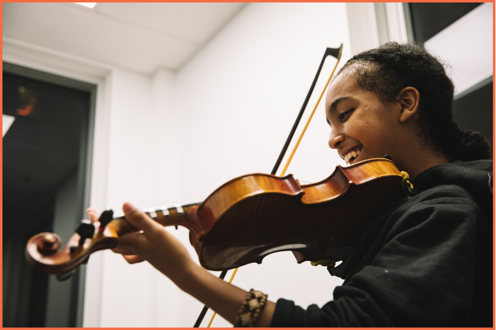 A young girl with curly hair smiles while playing the violin in a music room, holding the bow in her right hand and the violin in her left.