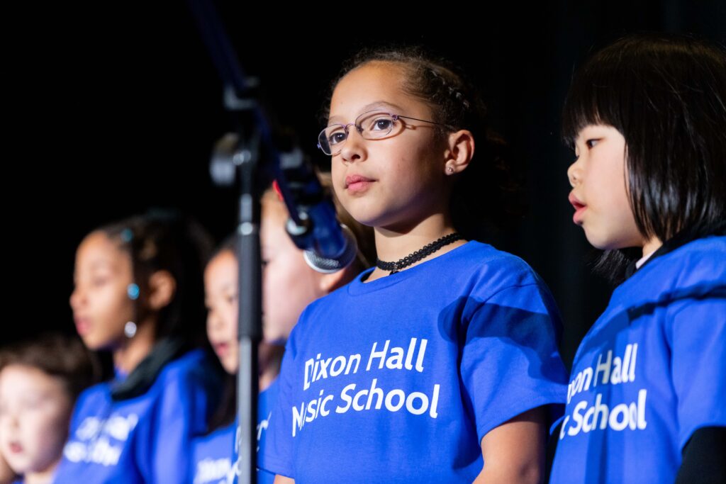 A group of children in blue "Dixon Hall Music School" t-shirts sing into microphones on stage, with a young girl wearing glasses standing in focus.