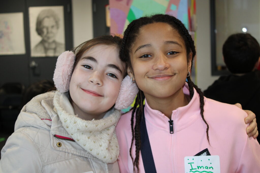 Two young girls smile and pose together indoors. One wears a beige coat with pink earmuffs, while the other wears a light pink jacket. They stand close with their arms around each other, with a classroom setting in the background.