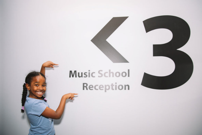 A young girl with braided hair and a bright smile points excitedly at a wall sign that reads "Music School Reception" with a large "<3" symbol above it.