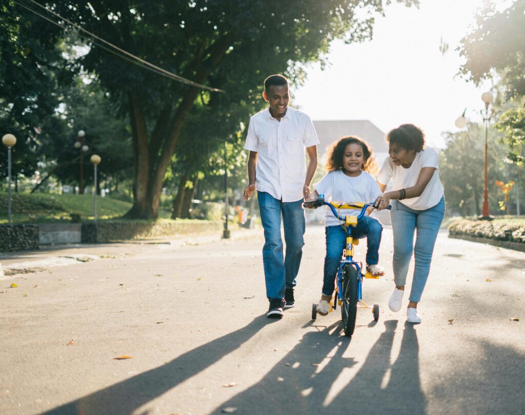A young girl with curly hair is learning to ride a yellow and blue bicycle with training wheels on a tree-lined path. Her mother, dressed in a white shirt and jeans, is holding the bike and smiling, while her father, also in a white shirt and jeans, walks alongside with an encouraging expression. The setting is a peaceful park with greenery, lampposts, and soft sunlight filtering through the trees.