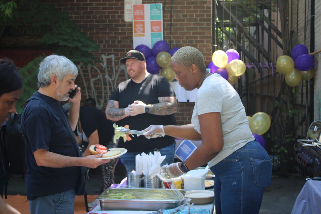 a-woman-wearing-gloves-is-serving-food-to-a-man-holding-a-plate-while-talking-on-his-phone-another-man-stands-behind-them-smiling-the-setting-is-an-outdoor-event-with-purple-and-gold-balloons-a-brick-wall-with-posters-and-a-buffet-table-with-various-food-items