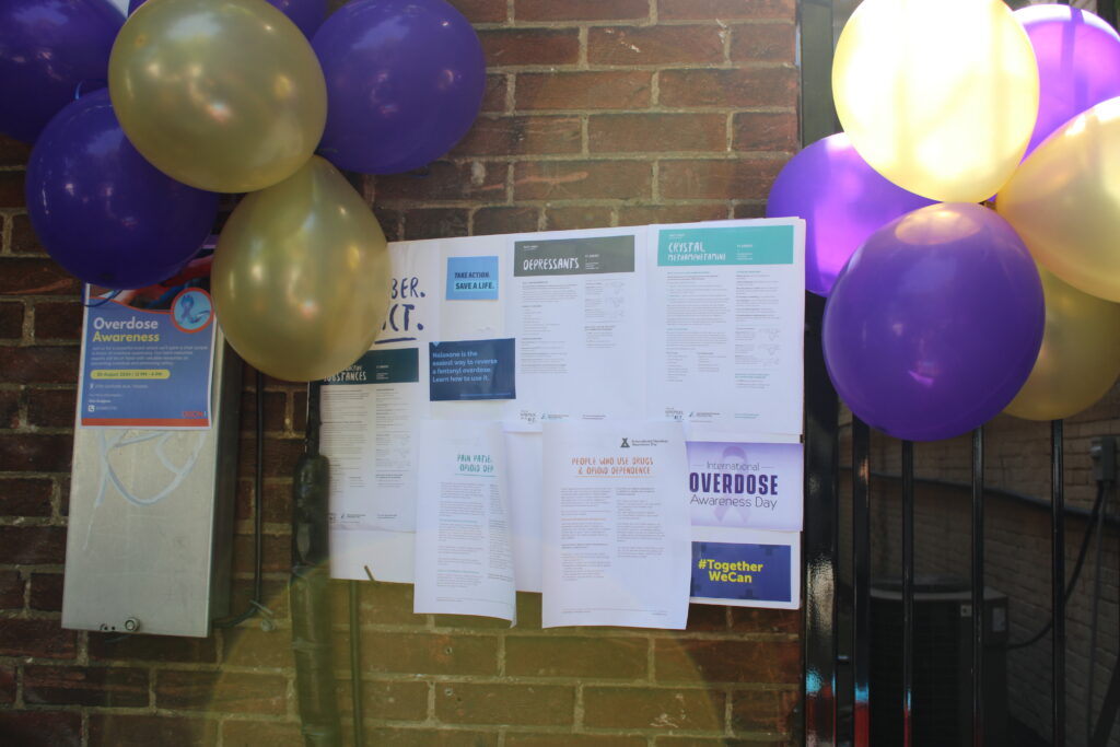 a-brick-wall-is-decorated-with-purple-and-gold-balloons-and-covered-with-informational-posters-about-overdose-awareness-and-harm-reduction-some-of-the-posters-include-details-on-substance-use-and-support-resources
