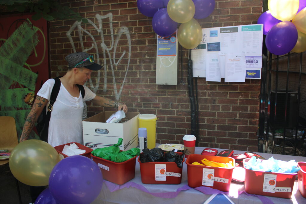 a-person-wearing-a-white-shirt-and-a-cap-is-reaching-into-a-storage-box-at-an-outdoor-table-decorated-with-purple-and-gold-balloons-the-table-holds-labeled-bins-with-supplies-a-coffee-cup-and-harm-reduction-kits-a-brick-wall-with-posters-and-graffiti-is-in-the-background