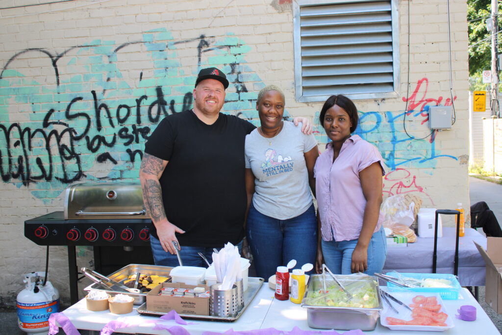 three-people-are-standing-together-outdoors-smiling-at-the-camera-behind-them-a-table-decorated-with-purple-and-gold-balloons-holds-supplies-and-a-brick-wall-with-posters-is-visible-a-graffiti-covered-van-is-parked-nearby