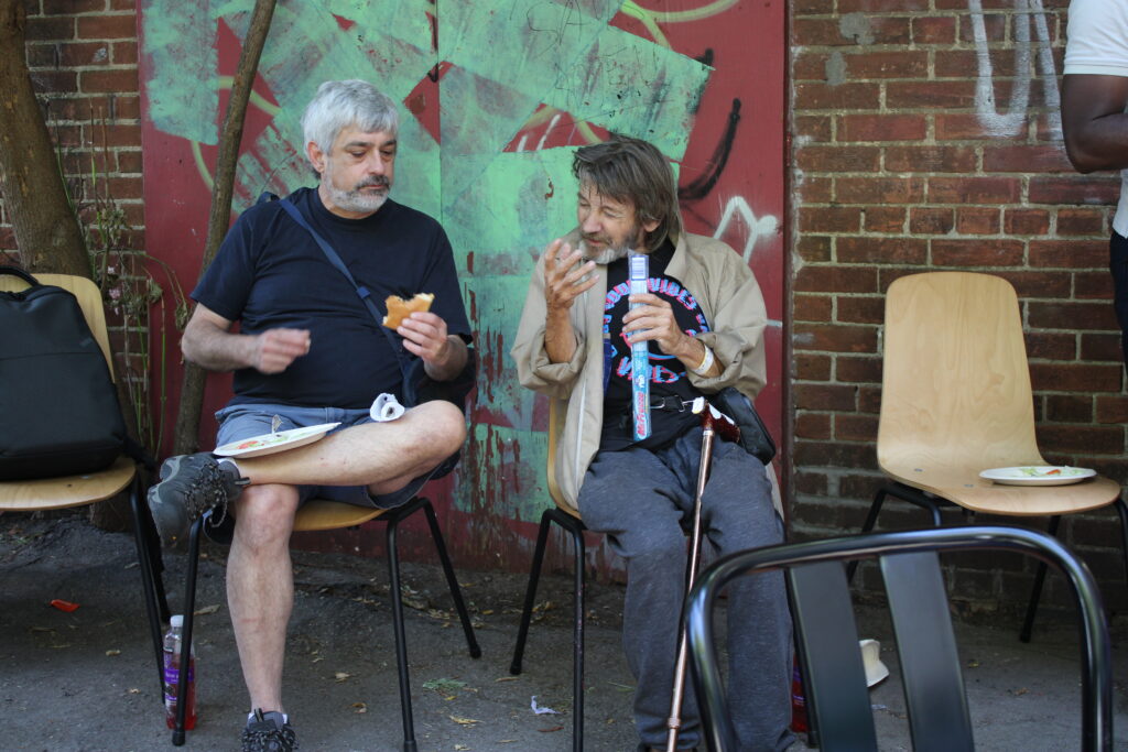 two-men-are-seated-outdoors-on-chairs-having-a-conversation-while-eating-one-holds-a-sandwich-and-the-other-has-a-walking-cane-and-a-snack-a-graffiti-covered-wall-is-behind-them-along-with-other-empty-chairs