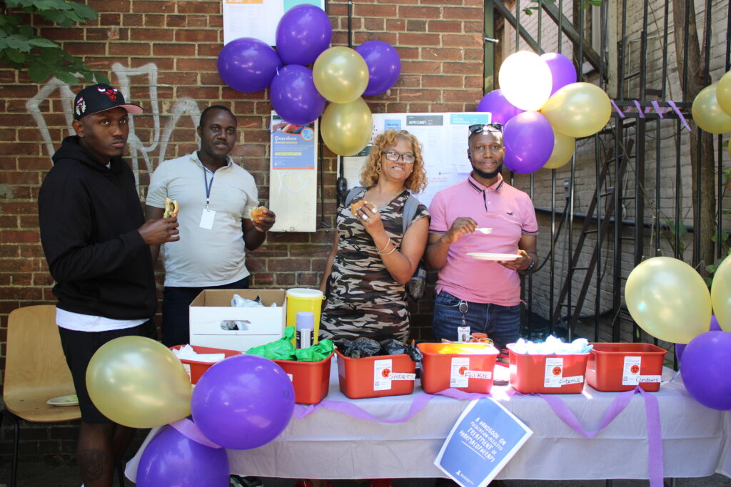 four-people-are-gathered-around-an-outdoor-table-decorated-with-purple-and-gold-balloons-they-are-holding-food-and-drinks-while-standing-behind-bins-labeled-with-different-supplies-a-brick-wall-with-posters-and-notices-is-in-the-background