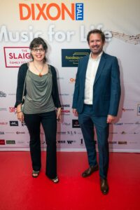 A smiling man and woman pose on the red carpet at the "Music for Life" event, standing in front of a branded backdrop featuring sponsor logos.