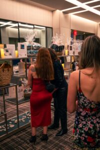 Guests browse a silent auction display featuring gift baskets, framed pictures, and various items wrapped in cellophane. The display is illuminated by small candles, and attendees in elegant attire examine the auction details.