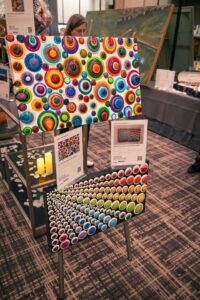 Two colorful abstract paintings featuring vibrant circular patterns are displayed on a stand at an event. Informational cards with details about the artwork are placed below each painting. The background shows a venue with decorative elements and auction items.