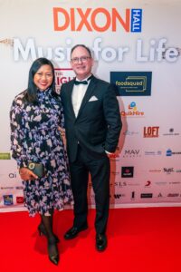 A well-dressed couple poses on the red carpet at the "Music for Life" event, standing in front of a backdrop featuring sponsor logos and event branding.