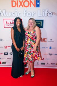 Two women posing and smiling on the red carpet at the "Music for Life" event, with a branded backdrop featuring sponsor logos behind them. One woman is wearing a black dress and holding a black purse, while the other is in a colorful floral dress with teal heels.