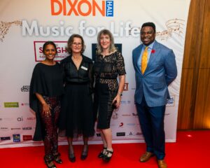 Four elegantly dressed individuals pose on the red carpet in front of a "Music for Life" event backdrop featuring sponsor logos. The group includes three women in stylish black outfits and a man in a blue suit with an orange tie, all smiling for the camera.