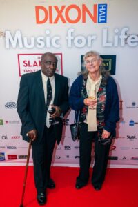 An older man and woman pose together on the red carpet at a "Music for Life" event, holding glasses of wine. The man wears a suit and tie while holding a cane, and the woman wears a patterned scarf and cardigan. The event backdrop features sponsor logos.